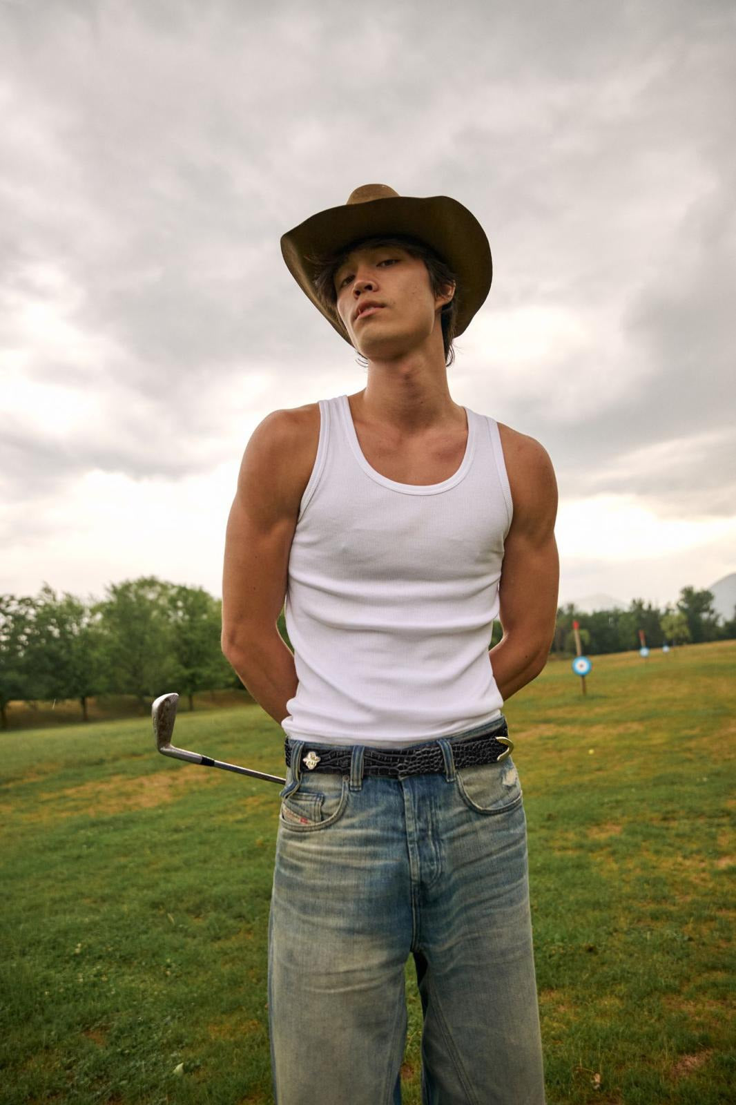 Sergi wearing a white tank top, blue jeans, black belt and a cowboy hat standing on a grassy field with a cloudy sky.