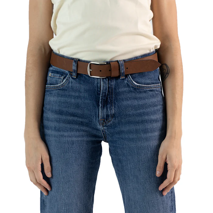 En la foto aparece la cadera de una persona que lleva un cinturon clasico de cuero de bufalo marron y hebilla metálica sujetando unos jeans azules y una camiseta blanca.