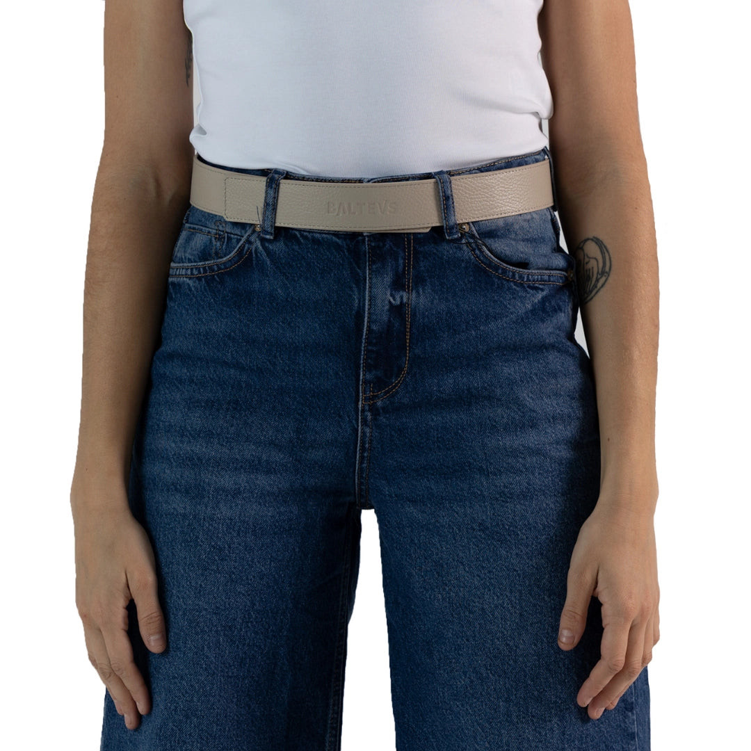 En la foto aparece la cadera de una persona que lleva un cinturon de cuero gris piedra con cierre de velcro sujetando unos jeans azules y una camiseta blanca.