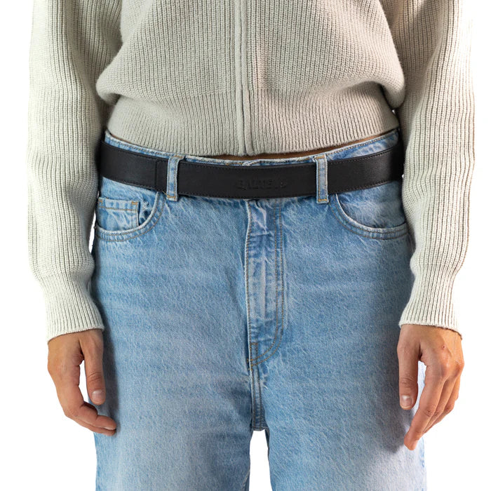 En la foto aparece la cadera de una persona que lleva un cinturon de cuero negro con cierre de velcro sujetando unos jeans azules y un jersey gris.