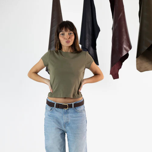 En la foto aparece una chica que lleva un cinturon clasico de cuero de bufalo y hebilla dorada en forma de serpiente, sujetando unos jeans y una camiseta verde. De fondo, cuelgan mantas de cuero de distintos colores.