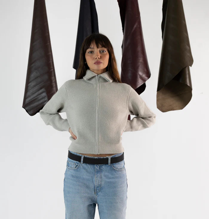 En la foto aparece una chica que lleva un cinturon de cuero negro y cierre de velcro, sujetando unos jeans y un jersey gris. De fondo, cuelgan mantas de cuero de distintos colores.
