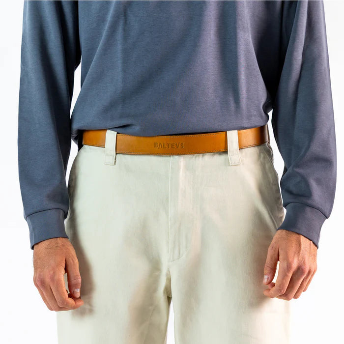 En la foto aparece la cadera de una persona que lleva un cinturon de cuero color mostaza con cierre de velcro sujetando unos pantalones blancos y una camiseta azul.