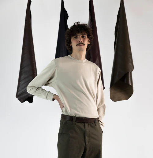 En la foto aparece un chico que lleva un cinturon de cuero de bufalo marron y cierre de velcro, sujetando un pantalon verde y una camiseta gris. De fondo, cuelgan mantas de cuero de distintos colores.