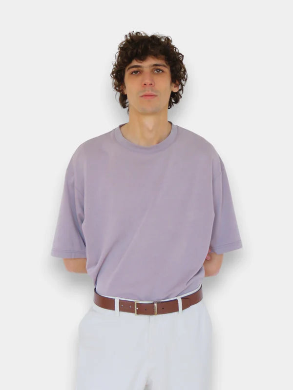 En la foto aparece una chico que lleva un cinturon de color marron con hebilla metálica reversible, sujetando un pantalon blanco y una camiseta lila.