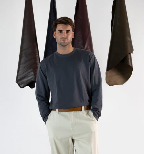 En la foto aparece un chico que lleva un cinturon de cuero mostaza y cierre de velcro, sujetando unos pantalones blancos y una camiseta azul. De fondo, cuelgan mantas de cuero de distintos colores.